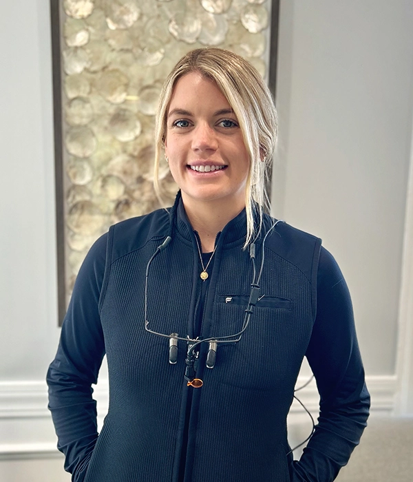 Dentist Kaeleigh Morton In Vest Smiling In Front Of Grey Wall